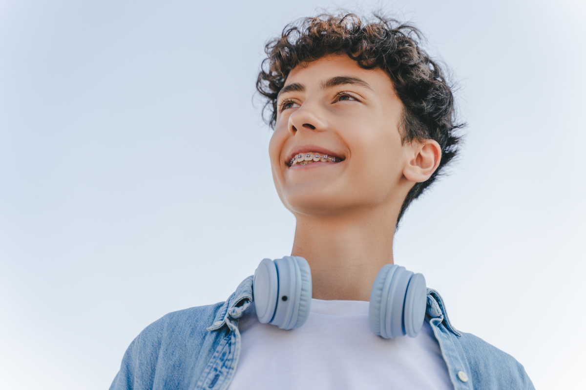 Portrait of smiling curly haired teenager with braces wearing headphones looking away standing on the street. Inspiration, technology concept