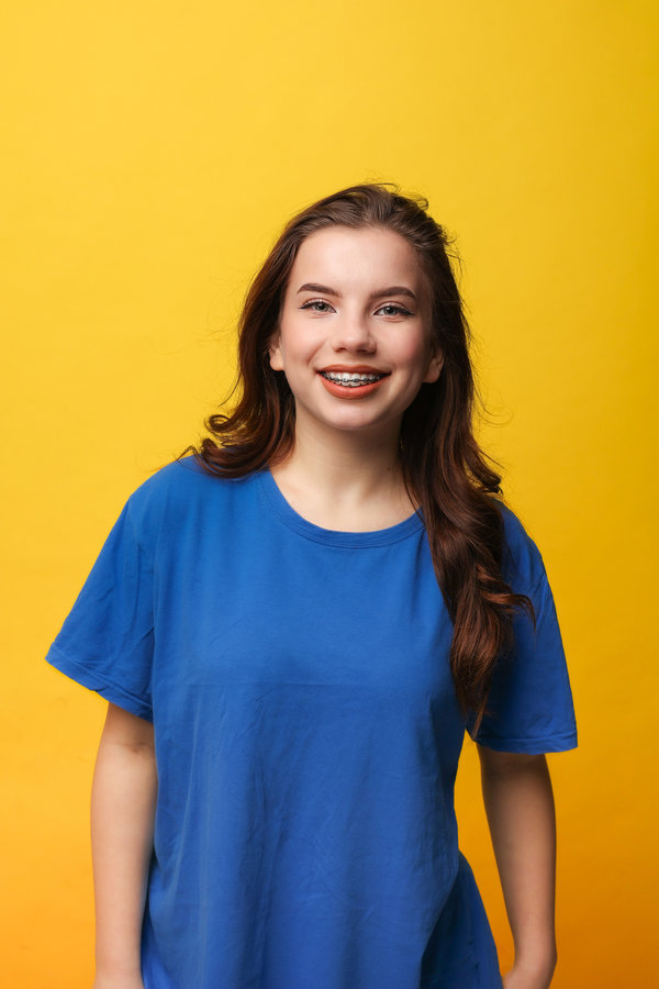 Positive teen girl with dental braces in blue t-shirt. Hands behind head, wide smile, looks at camera on yellow backdrop. Dental care services, orthodontic clinics, teen health campaigns, youth