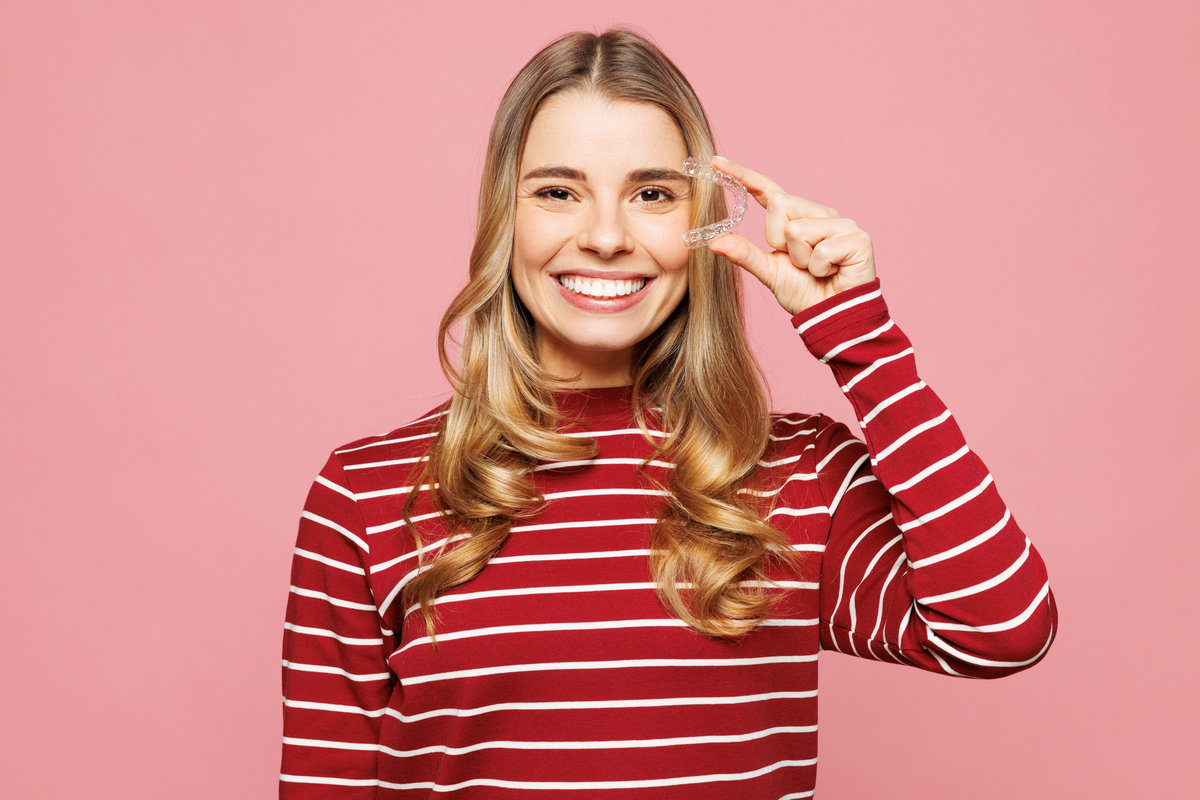 Young smiling woman wear red striped sweatshirt casual clothes hold in hand cover eye with invisible transparent aligners, invisalign dental braces isolated on plain pink background. Lifestyle concept