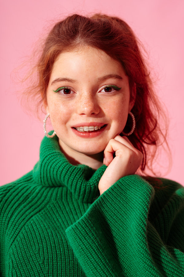 Happy smiling freckled redhead teenager girl with dental retainer, wearing trendy green knitted turtleneck sweater, posing on pink background