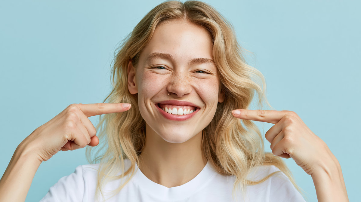 Young woman with freckles happily pointing to her perfect white smile against a blue background